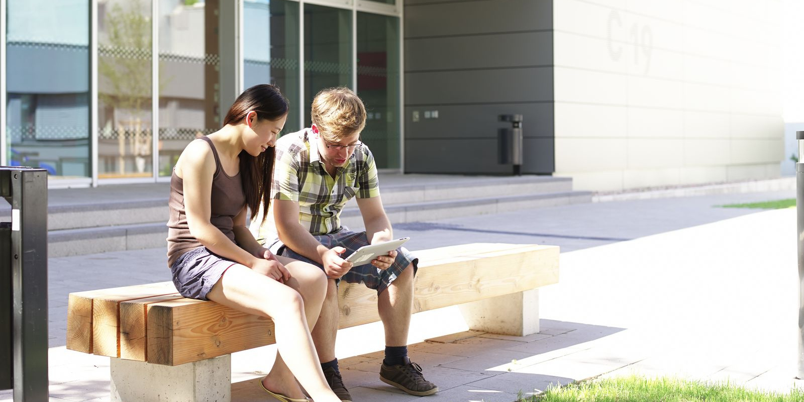 Zwei Studierende sitzen auf einer Bank und betrachten ein Tablet.